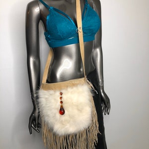 May include: Cream-colored, fringed crossbody bag with a white fur front. The bag features a tan leather strap and a decorative pendant with amber-colored beads. The mannequin is wearing a teal bra.