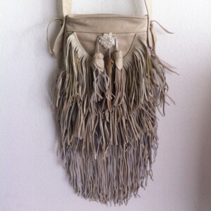 May include: A beige leather crossbody bag with fringe detailing. The bag has a beaded embellishment on the front.