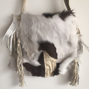 May include: A white and brown cowhide bag with fringe details. The bag is made of soft, supple leather and has a unique, rustic look.