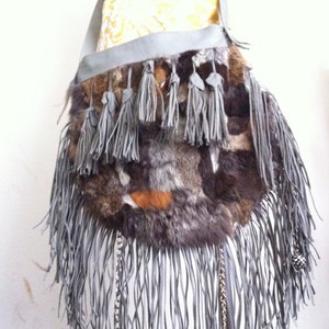 May include: A gray faux fur crossbody bag with a long fringe and silver chain accents. The bag has a flap closure and a strap that can be adjusted.