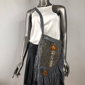 May include: A gray fringed crossbody bag with a brown leather flap and amber colored accents. The bag is worn on a mannequin.