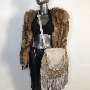 May include: A gray leather crossbody bag with fringe details. The bag is made of a soft, supple leather and has a unique, bohemian style. The bag is worn by a mannequin wearing a brown fur jacket and black pants.