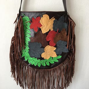 May include: A brown leather shoulder bag with a fringe trim. The bag is decorated with colorful leather leaves in shades of green, red, yellow, and brown.