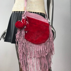 May include: A red and pink suede fringe bag with a long strap. The bag has a round shape and is decorated with a small red fur pom-pom.