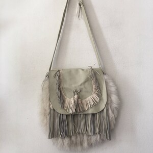 May include: A light beige leather crossbody bag with fringe detailing and a faux fur trim. The bag has a long strap and a flap closure.