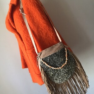 May include: A brown suede fringe bag with a gray sheepskin lining and a beaded accent. The bag is hanging on a bright orange coat.