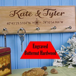 Op de afbeelding: Een houten sleutelhouder gegraveerd met de namen "Kate & Tyler" en co&ouml;rdinaten. Het heeft vier zilveren sleutelhaken. De tekst "Engraved Butternut Hardwood" is ook zichtbaar.