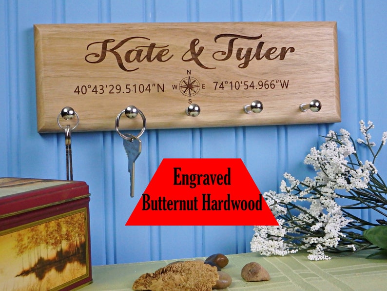 Op de afbeelding: Een houten sleutelhouder gegraveerd met de namen "Kate & Tyler" en co&ouml;rdinaten. Het heeft vier zilveren sleutelhaken. De tekst "Engraved Butternut Hardwood" is ook zichtbaar.