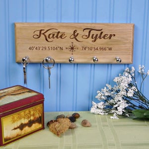 Op de afbeelding: Een houten sleutelhouder met de namen "Kate & Tyler" gegraveerd, co&ouml;rdinaten en een kompasroos. Het heeft vijf zilveren sleutelhaken. Een decoratieve blik en sleutels zijn zichtbaar.