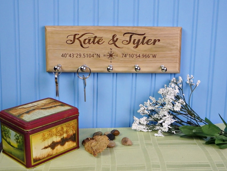 Op de afbeelding: Een houten sleutelhouder met de namen "Kate & Tyler" gegraveerd, co&ouml;rdinaten en een kompasroos. Het heeft vijf zilveren sleutelhaken. Een decoratieve blik en sleutels zijn zichtbaar.