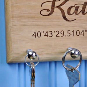 Op de afbeelding: Houten sleutelhouder met de naam "Kate" gegraveerd in een sierlijk lettertype. De sleutelhouder heeft ook gegraveerde co&ouml;rdinaten en twee zilveren sleutelringen met sleutels eraan. De achtergrond is lichtblauw.