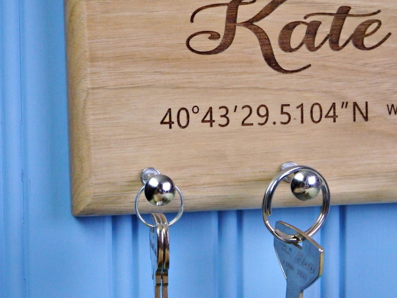 Op de afbeelding: Houten sleutelhouder met de naam "Kate" gegraveerd in een sierlijk lettertype. De sleutelhouder heeft ook gegraveerde co&ouml;rdinaten en twee zilveren sleutelringen met sleutels eraan. De achtergrond is lichtblauw.