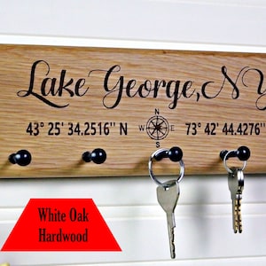 Op de afbeelding: Een houten sleutelhouder met de tekst "Lake George, NY" en co&ouml;rdinaten. Het heeft vier zwarte knoppen om sleutels op te hangen. Gemaakt van wit eiken hardhout.