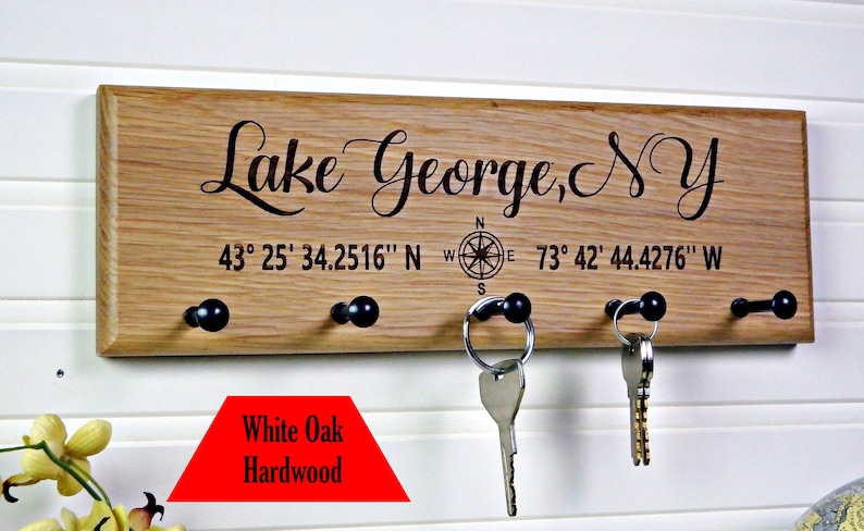 Op de afbeelding: Een houten sleutelhouder met de tekst "Lake George, NY" en co&ouml;rdinaten. Het heeft vier zwarte knoppen om sleutels op te hangen. Gemaakt van wit eiken hardhout.