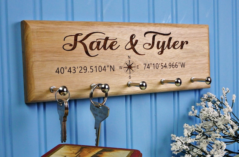 Personalized Light Tan/Brown Butternut wood key holder, 4"x12", with 5 chrome pegs, laser engraved script text "Kate & Tyler", compass graphic and their GPS coordinates shown mounted on a light blue wall with decorative flowers in the background.