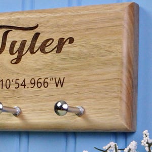 Op de afbeelding: Houten sleutelhouder met de naam "Tyler" gegraveerd in een cursief lettertype. De sleutelhouder heeft twee zilverkleurige haken en co&ouml;rdinaten gegraveerd onder de naam. Het hout heeft een natuurlijke, lichtbruine kleur.
