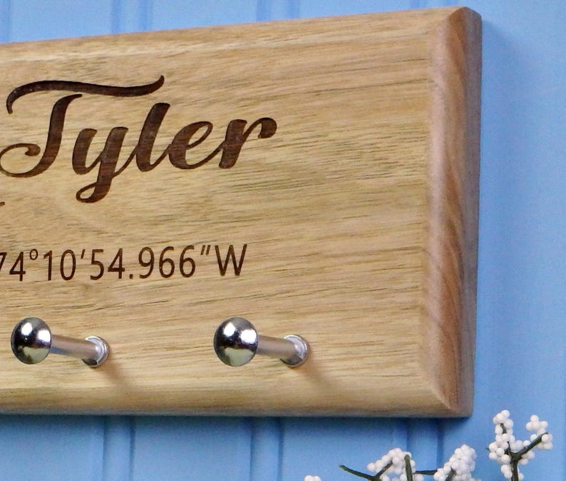 Op de afbeelding: Houten sleutelhouder met de naam "Tyler" gegraveerd in een cursief lettertype. De sleutelhouder heeft twee zilverkleurige haken en co&ouml;rdinaten gegraveerd onder de naam. Het hout heeft een natuurlijke, lichtbruine kleur.