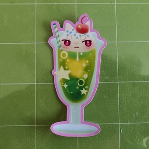 Puede incluir: Una pegatina de un flotador de soda verde y amarillo con una cara de gato blanco en la parte superior. El gato tiene ojos rojos y una nariz rosa. El flotador de soda tiene una cereza en la parte superior y una pajita.