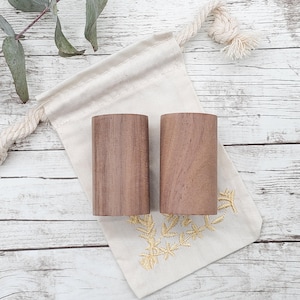 May include: Two wooden cylinders in a natural brown color, sitting on a white fabric bag with gold embroidery. The bag has a drawstring closure.