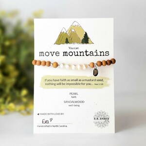 May include: A wooden bracelet with white pearls and a silver charm that says "You can move mountains". The bracelet is on a white card with a green mountain illustration and the text "If you have faith as small as a mustard seed, nothing will be impossible for you... Mat 17:20". The card also says "PEARL faith SANDALWOOD well-being" and "MADE WITH LOVE BY: Eva".