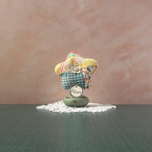 May include: A decorative star-shaped object made of fabric with floral and plaid patterns. It sits atop a green base and a white lace doily. A small tag with the word "bloom" hangs from the star.