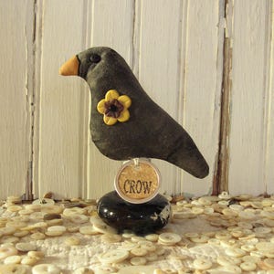 May include: A handcrafted, dark gray crow figurine with a yellow and brown flower accent. The crow has a yellow beak and is perched on a black, round base. A tag with the word "CROW" is attached to the base.