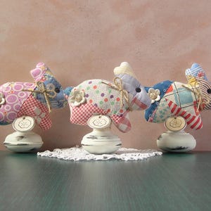 May include: Three decorative fabric bunny figurines in patchwork patterns of pink, blue, and white, each mounted on a white, distressed pedestal. The bunnies have flower accents and small tags.