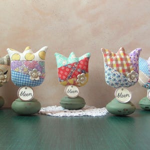 May include: Five fabric tulip decorations with various patterns and colors, including floral, plaid, and polka dots. Each tulip has a small flower embellishment and a tag that says "bloom". They are set on green, rounded bases.