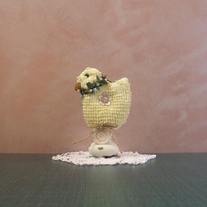 May include: A handmade, yellow crochet chick figurine with a floral wreath and a flower embellishment. The chick sits atop a white base with a decorative tag, resting on a small, white lace doily. The background is a muted pink.