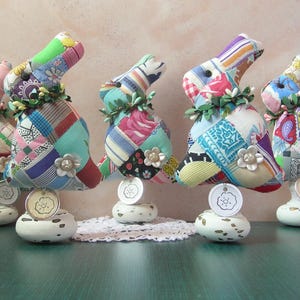 May include: A collection of colorful, patchwork fabric bunny figurines. Each bunny has a floral garland around its neck and a decorative flower accent. They stand on white, distressed pedestals with small tags.