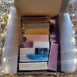 May include: A box filled with a variety of handmade soap bars. The bars are arranged in a stack and include colours like brown, green, yellow, pink, white, blue, and purple.