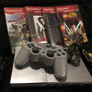 May include: A silver PlayStation 2 console with a gray controller, a black cable, and a black and silver device with the text "AUTO RF SWITCH" on it. The console is sitting on a black surface. There are five video game cases in the background, including "ATV Offroad Fury", "Max Payne", "Spider-Man", "Lego Star Wars: The Video Game", and another game with a black cover.