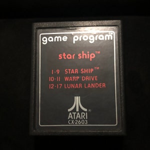 Puede incluir: Cartucho de juego Atari negro con una etiqueta roja que dice "game program star ship" y enumera los juegos "STAR SHIP", "WARP DRIVE" y "LUNAR LANDER". La etiqueta también incluye el logotipo de Atari y el texto "ATARI CX-2603".