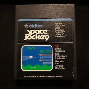 Puede incluir: Un cartucho de videojuego Space Jockey negro. La etiqueta presenta el logotipo de Vidtec y el título del juego en blanco. El gráfico del juego muestra un cielo azul con objetos blancos y naranjas. El texto de la etiqueta incluye "16 variaciones de juego".