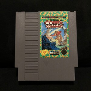 May include: A vintage Nintendo Entertainment System game cartridge for Cobra Command, a Data East arcade game. The cartridge is gray with a green, yellow, and red label featuring the game title and a pixelated image of a helicopter.