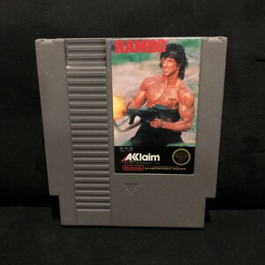 Op de afbeelding: Een grijze Nintendo Entertainment System-spelcartridge met een label met een foto van Sylvester Stallone als John Rambo uit de film "Rambo: First Blood Part II". Het label bevat ook de titel "RAMBO" in rode letters en het logo van de game-ontwikkelaar, "Acclaim Entertainment, Inc."