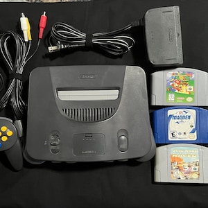Puede incluir: Una consola Nintendo 64 negra con un mando gris, cables AV y un adaptador de corriente. Hay tres cartuchos de juego: "Super Mario 64", "Madden NFL 99" y "Jet Force Gemini".