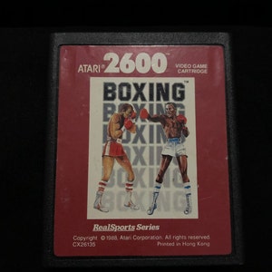 Può includere: Una cartuccia di videogioco Atari 2600 rossa e nera con il titolo "Boxing" e il testo "RealSports Series" stampato sul davanti. La cartuccia presenta una grafica di due pugili sul ring.