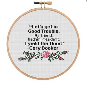 May include: A white embroidery hoop with a quote from Cory Booker: "Let's get in Good Trouble. My friend, Madam President, I yield the floor." The quote is surrounded by a floral design with pink roses and green leaves.