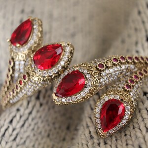 Puede incluir: Primer plano de joyería dorada adornada con grandes gemas rojas en forma de lágrima. Las joyas están incrustadas con piedras rojas y transparentes más pequeñas, sobre un fondo gris claro texturizado.