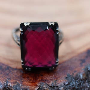 May include: A silver ring with a large, rectangular, faceted, deep red gemstone. The ring has a detailed band and is set against a wooden surface. The gemstone is the focal point.