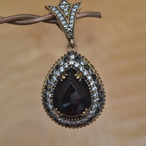 May include: A teardrop pendant featuring a large, faceted black stone. The stone is encircled by small, clear and black gemstones set in a gold-toned metal. The pendant hangs from a twisted metal chain, showcasing a vintage aesthetic.