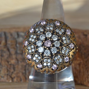 May include: A detailed, antique-style ring with a circular design. The ring features a gold-toned metal band with a floral pattern. The center is adorned with clear and pink gemstones, creating a sparkling effect. The ring is displayed on a clear stand.