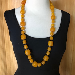 May include: A long, amber-colored beaded necklace displayed on a mannequin. The necklace features a variety of rounded and rectangular beads strung together with gold-colored wire. The beads vary in size and shape, creating a unique and eye-catching design.