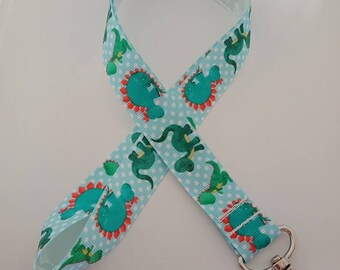 May include: A light blue lanyard with a repeating pattern of green dinosaurs and red accents. The lanyard has a white polka dot background and a silver-colored metal clasp.
