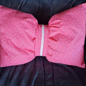Peut inclure: Coussin rose en forme de nœud papillon avec des pois blancs. Le coussin a une bande blanche au centre. Le coussin est sur une surface gris foncé.