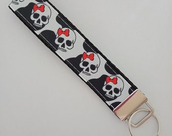 May include: A black and white key fob featuring a repeating pattern of skull faces with red bows. The key fob has a silver-colored metal clasp and a key ring.