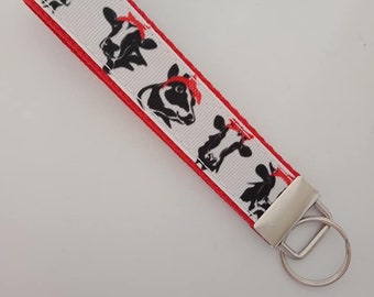 May include: A white key fob with a red border features a repeating pattern of black and white cow faces wearing red bandanas. The key fob has a silver metal key ring attached.