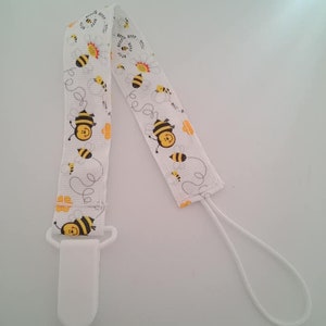 Op de afbeelding: Witte fopspeenclip met een bijenpatroon. De clip heeft een witte plastic clip en een wit lint met een herhalend patroon van gele en zwarte bijen, witte bloemen en een stippellijntje.