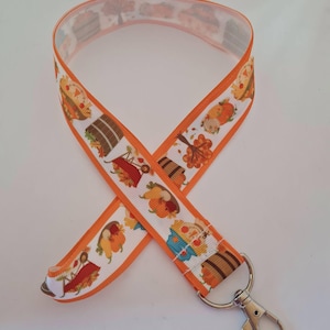May include: A white lanyard with orange edges featuring a repeating autumn-themed pattern of pumpkins, barrels, wheelbarrows, and fall foliage. The fabric strap is looped and attached to a silver-toned metal swivel lobster claw clasp.